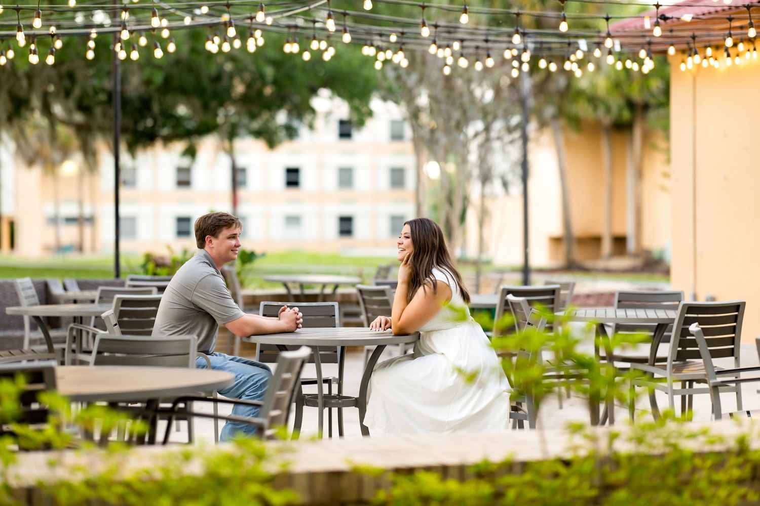 Couple enjoying outdoor patio dining under string lights surrounded by lush greenery and modern architecture.