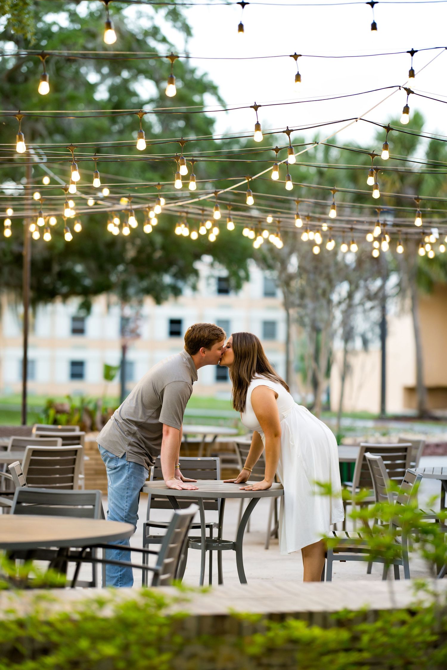 Couple sharing a romantic kiss across a table at an outdoor café adorned with warm string lights at dusk.