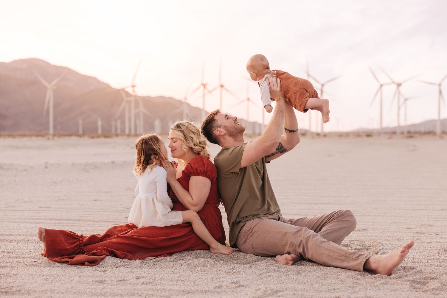 Family sitting on desert sand near wind turbines, parents holding baby up joyfully, golden hour light.