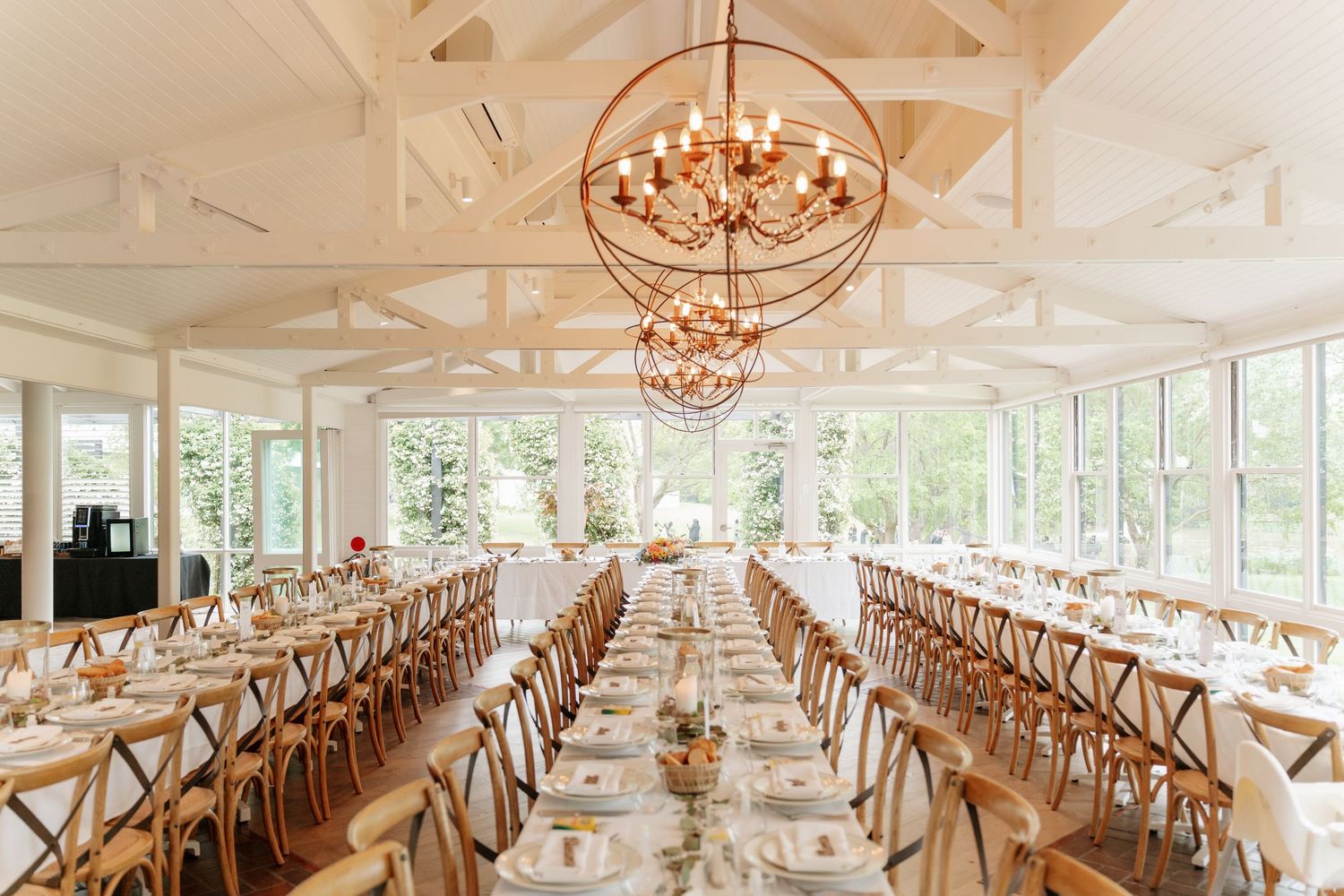 Elegant wedding reception hall with long tables, cross-back chairs, and a stunning gold orb chandelier in a bright conservatory.