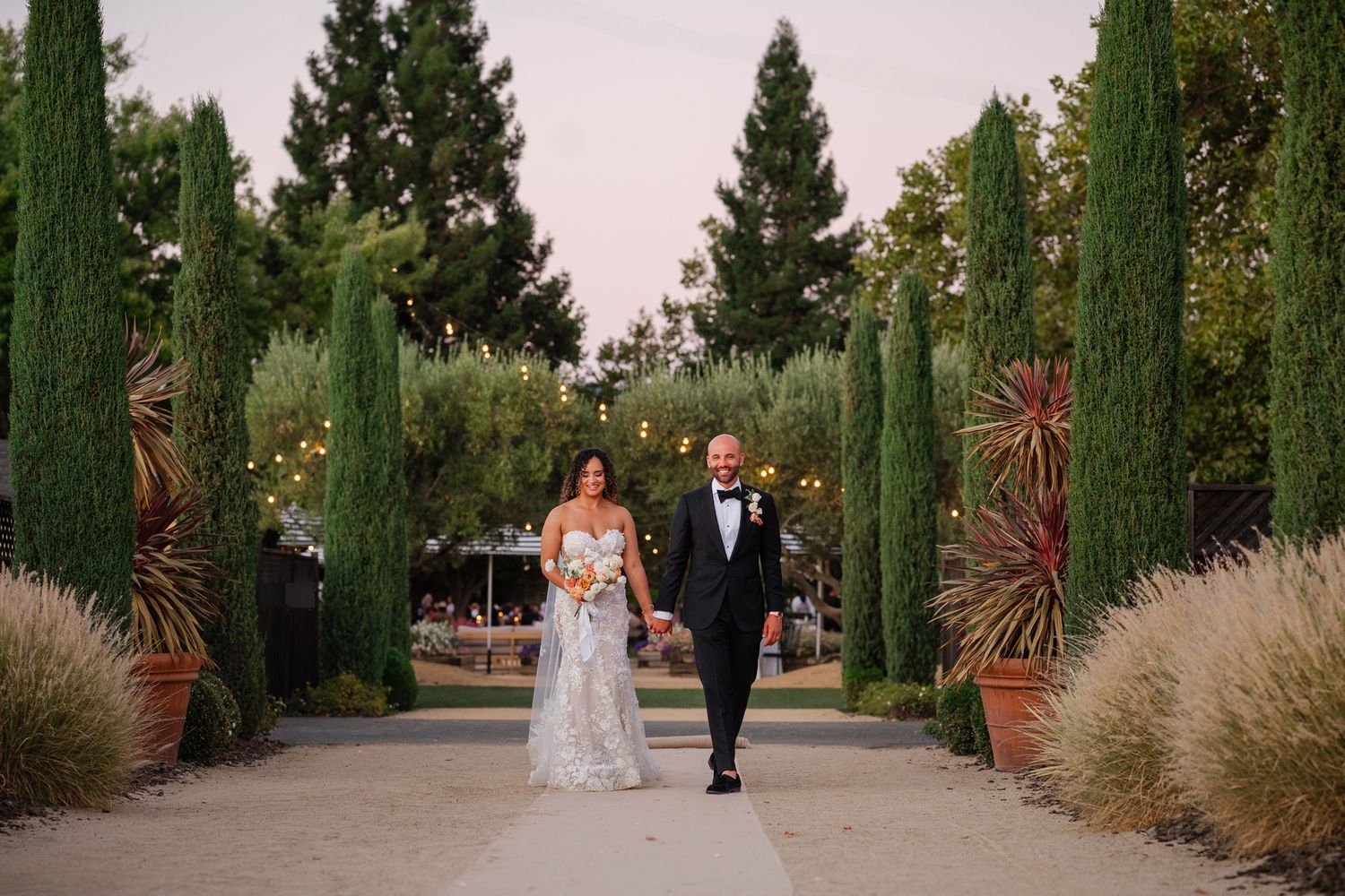 Bride and groom walking hand in hand down an elegant tree-lined garden path at dusk during outdoor wedding.