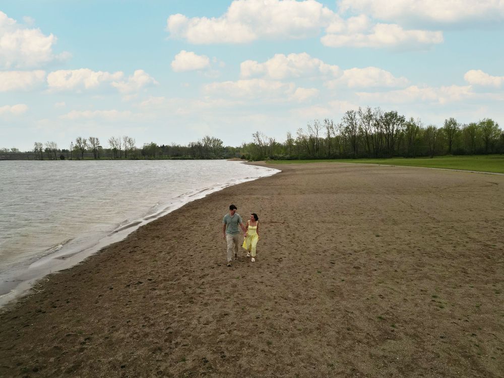 Couple holding hands walking along a sandy beach shoreline with trees and cloudy blue sky in the background.