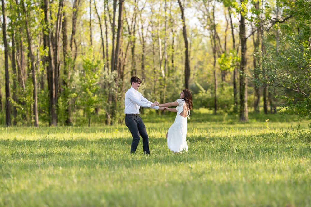 A couple dances joyfully in a lush green meadow surrounded by trees on a sunny day.