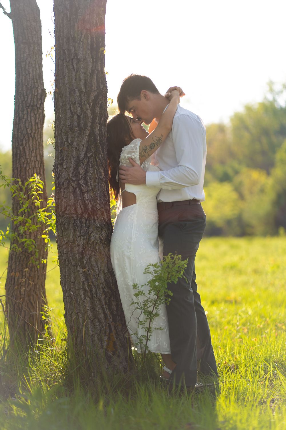 Couple sharing a romantic kiss leaning against a tree trunk in a sunlit green meadow, bride in white dress.