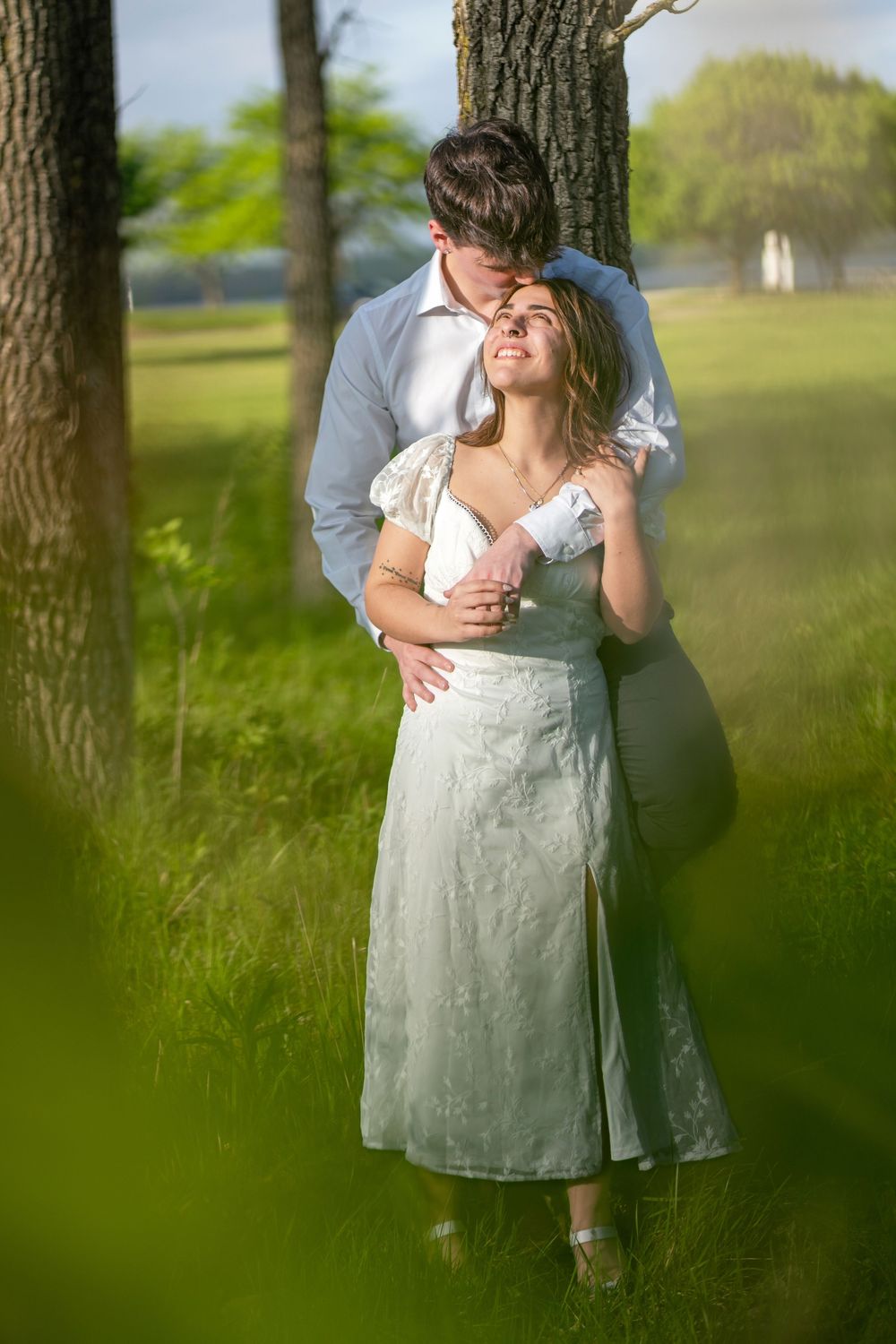 Romantic couple embracing near a tree in a lush green park, woman in white dress smiling warmly.