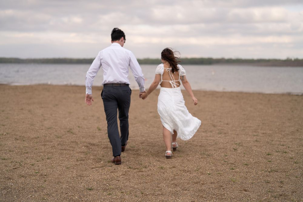 A couple in wedding attire walks hand-in-hand along a sandy beach shoreline on a cloudy day.