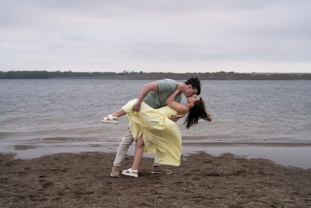 Romantic couple sharing a kiss on a sandy beach shore, man dipping woman in yellow dress by calm lake.