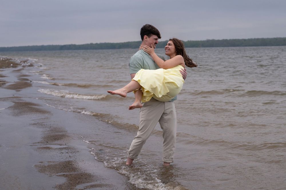 Romantic couple embracing on a beach shoreline, man carrying woman in yellow dress near ocean waves.