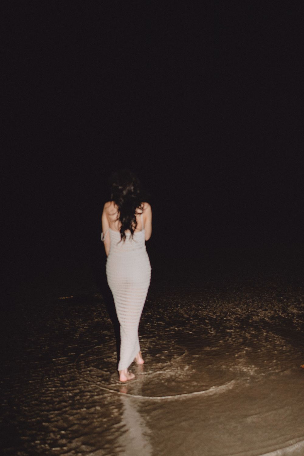 Woman in white dress walking barefoot on a rocky beach shore at night with waves washing around her feet.