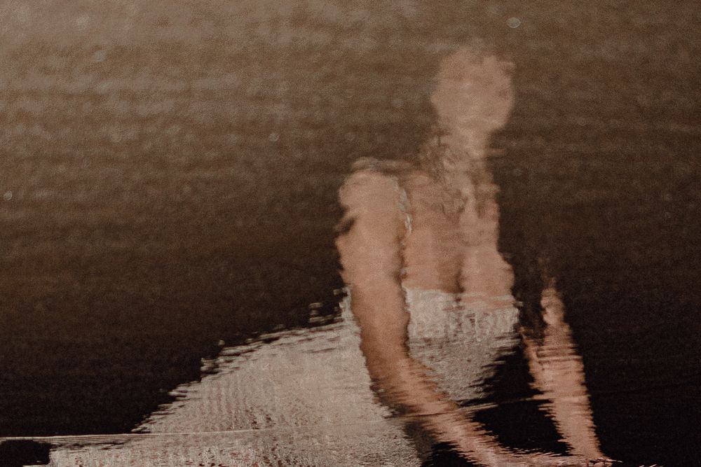 Blurry reflection of a person in white dress on dark water surface, creating an ethereal, ghostly abstract image.
