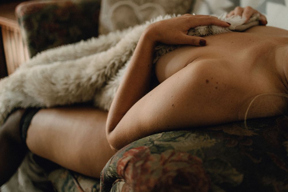 A nude figure resting on a floral armchair, partially draped with a soft fur blanket in warm, moody tones.