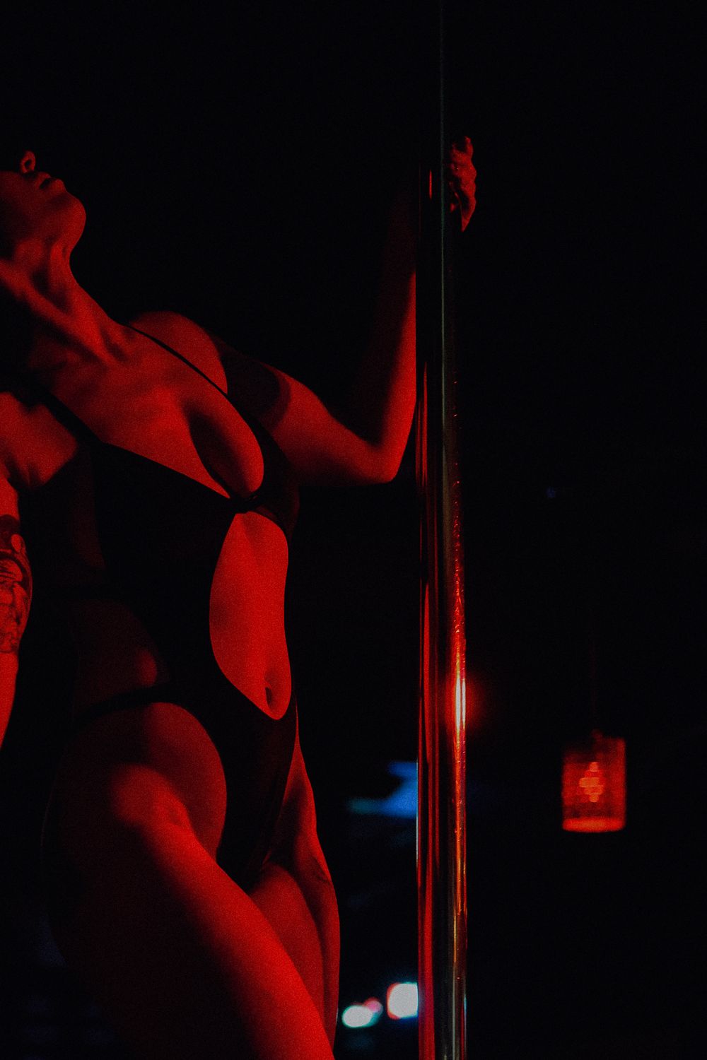 A dancer in a black cutout bodysuit poses against a pole under dramatic red lighting in a nightclub.