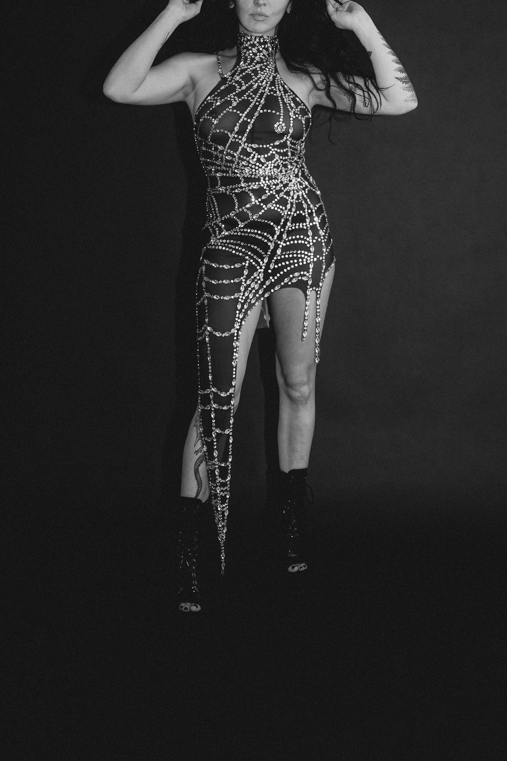 Woman posing in a rhinestone spider web halter dress with thigh-high slits and black boots, black and white photo.