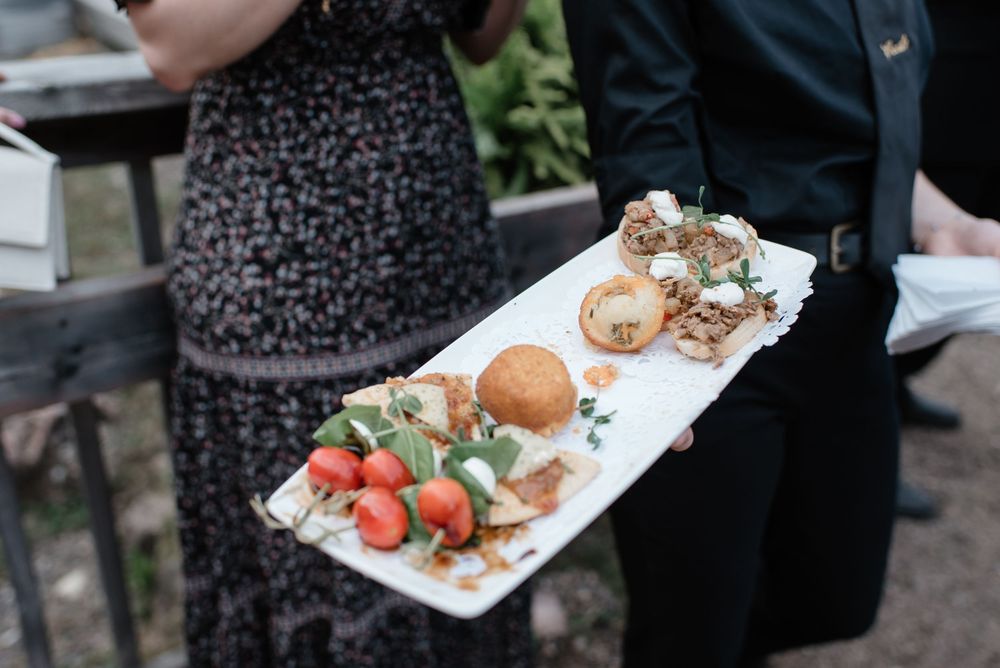 Person holding a white platter with elegant appetizers including strawberry skewers, a bread roll, and gourmet bites at an event.