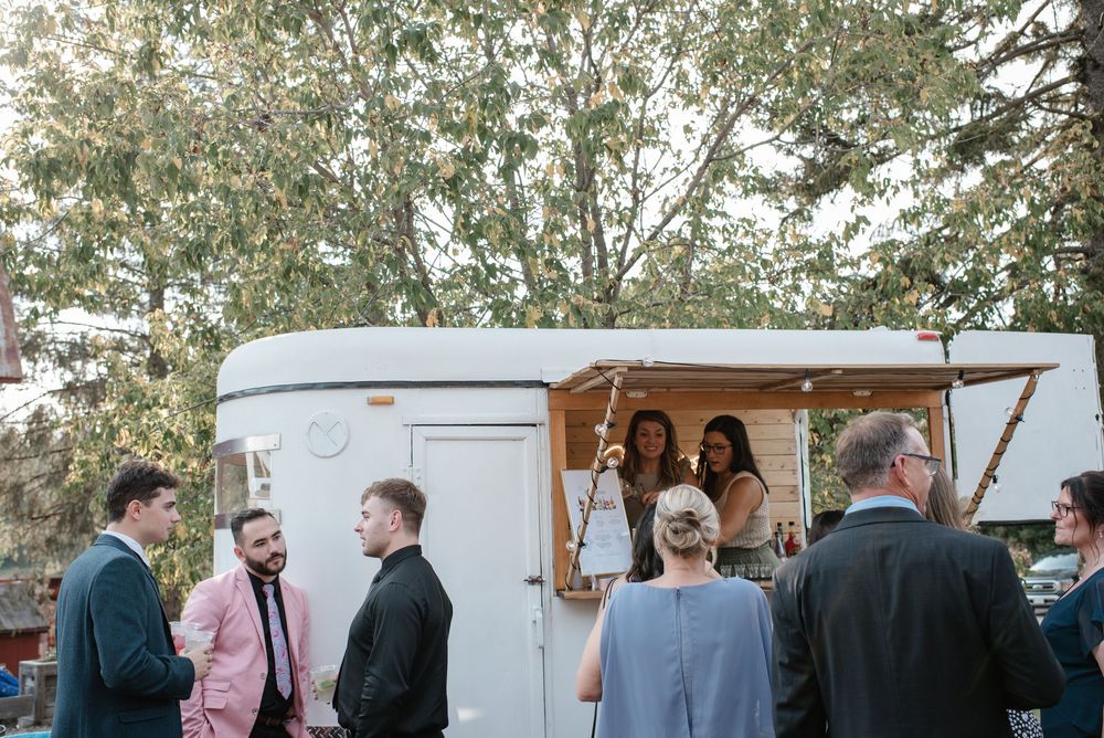 Guests mingle near a vintage white food trailer at an outdoor wedding reception surrounded by trees.