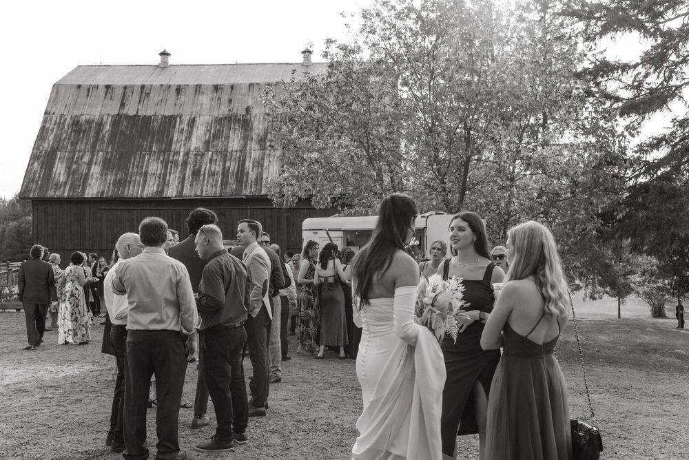 Black and white photo of wedding guests mingling outdoors near a rustic barn with blooming trees in background.
