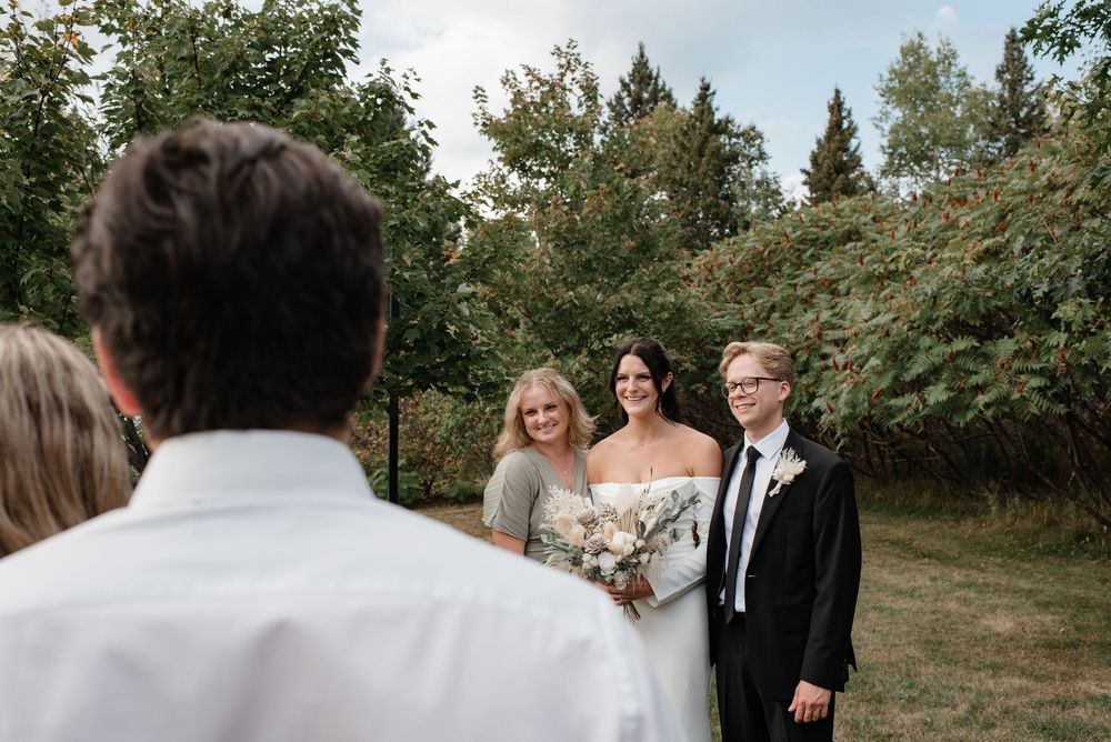 Bride in white gown holds bouquet beside two guests outdoors, with lush green trees in the background at a wedding.