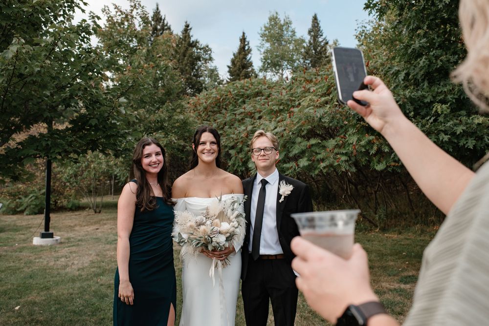 Wedding couple poses with guest outdoors while photographer captures the moment on a smartphone.