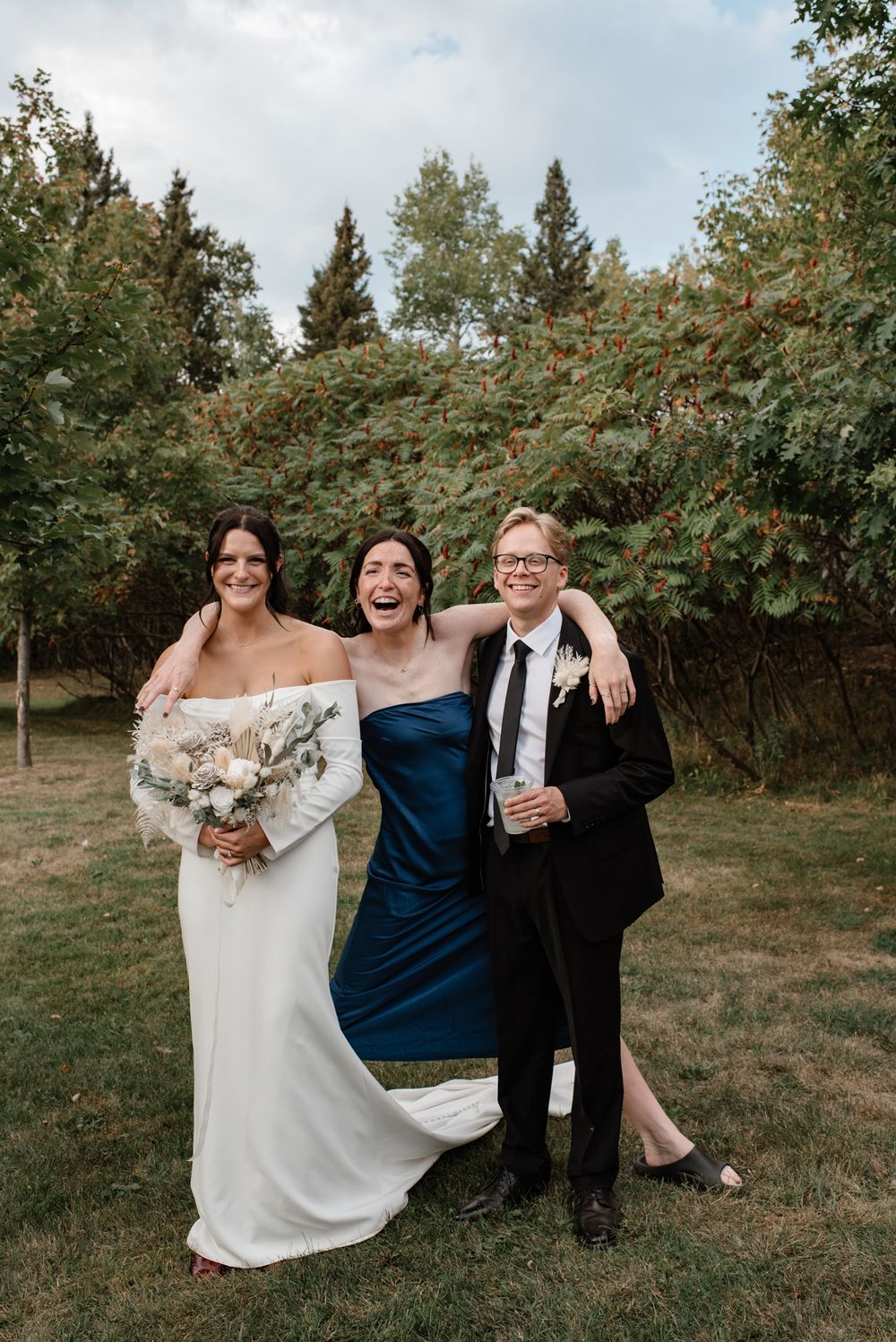 Bride in white gown poses with bridesmaid in blue dress and guest in black suit at outdoor wedding.
