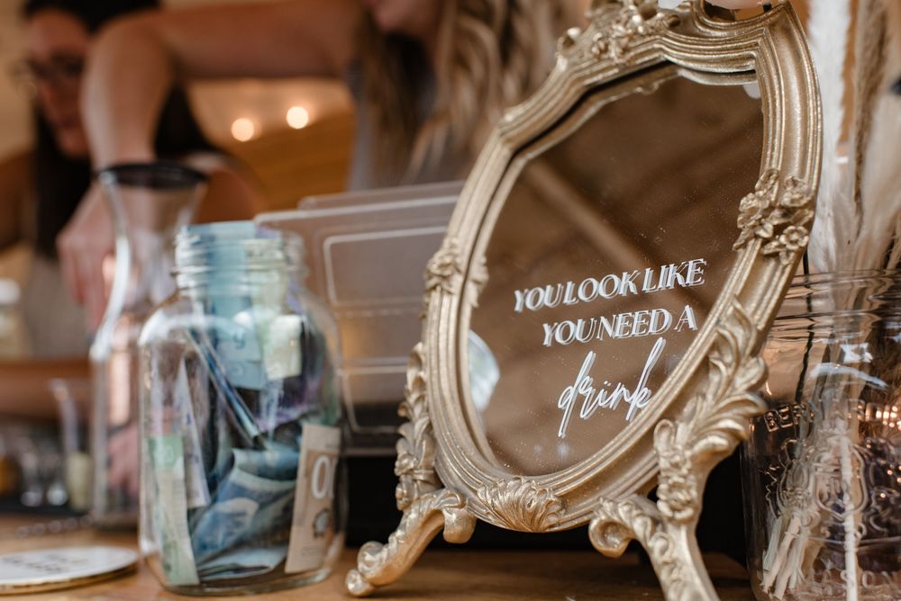 Ornate gold mirror with cursive text at a wedding bar setup, alongside glass jars filled with cash tips.