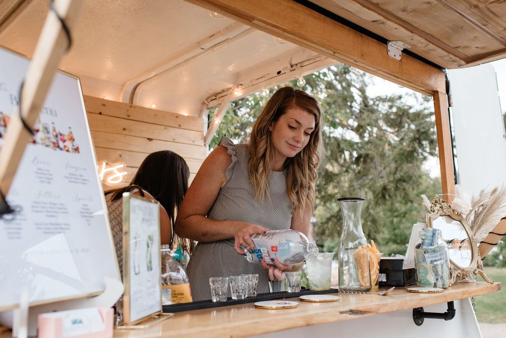 A woman pours a drink at a rustic wooden mobile bar decorated with string lights and glassware.