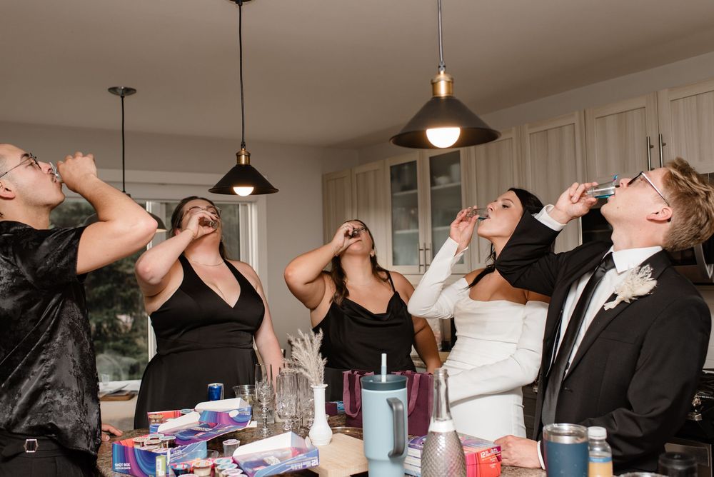 Bride and bridesmaids in black dresses taking shots together during wedding day preparations in a kitchen.