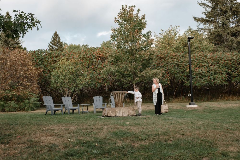 Two people stand on lush green lawn with Adirondack chairs and rustic backdrop at outdoor wedding venue.