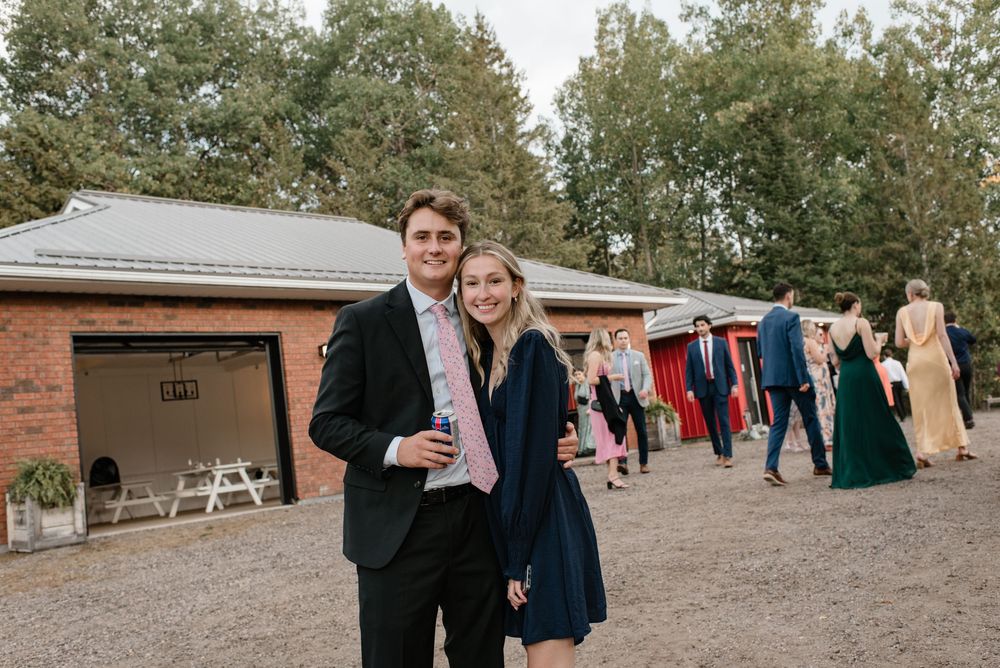 Couple smiling outdoors at a wedding venue, man in navy suit with pink tie, woman in blue dress.