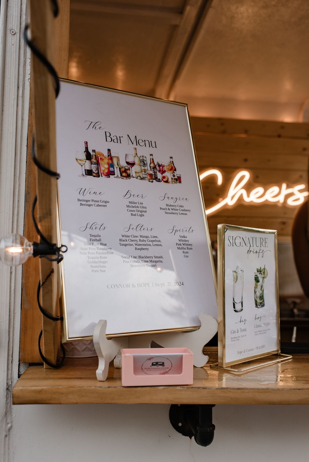 Elegant bar menu display with neon Cheers sign and signature drinks card at a wedding reception.