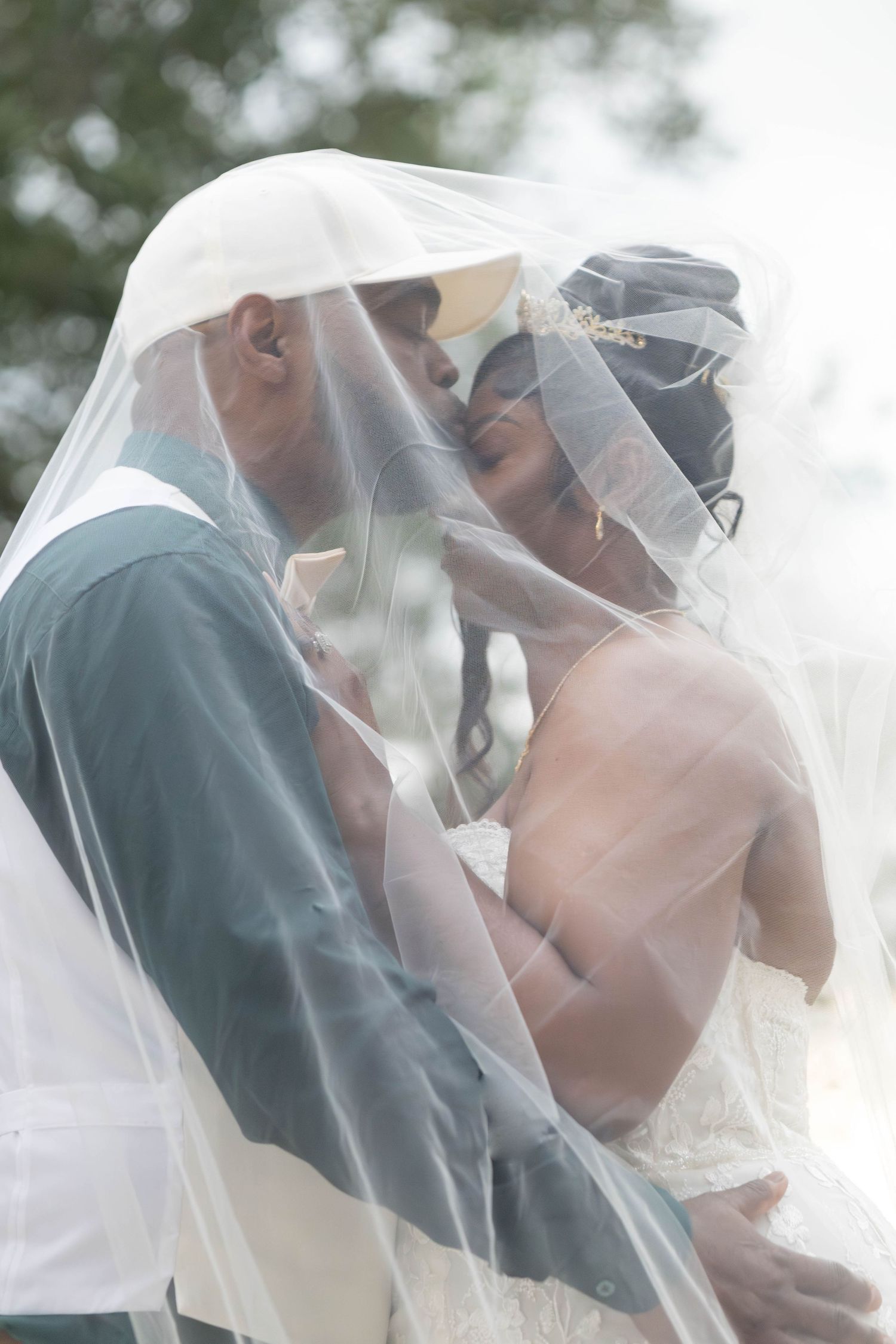 Bride and groom share a tender kiss under a flowing white veil outdoors.