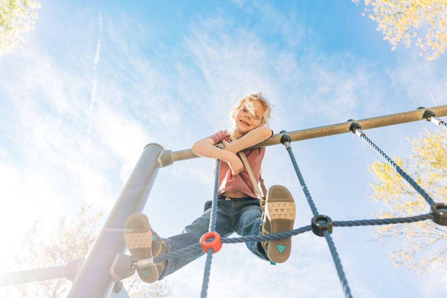Young boy climbing on playground rope structure under bright blue sky on sunny day.