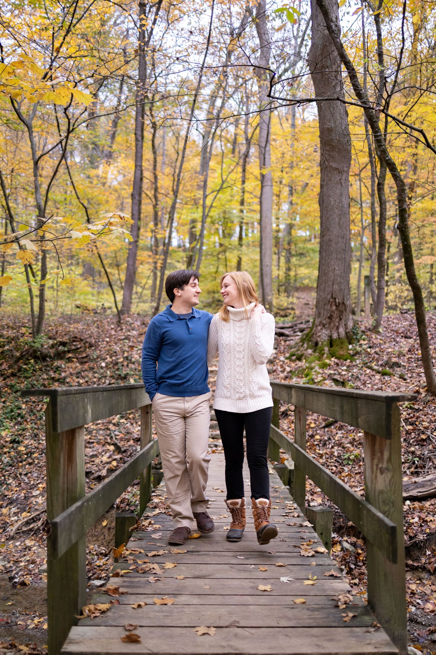 Couple walking hand-in-hand on a wooden bridge in Ault Park surrounded by colorful autumn foliage.