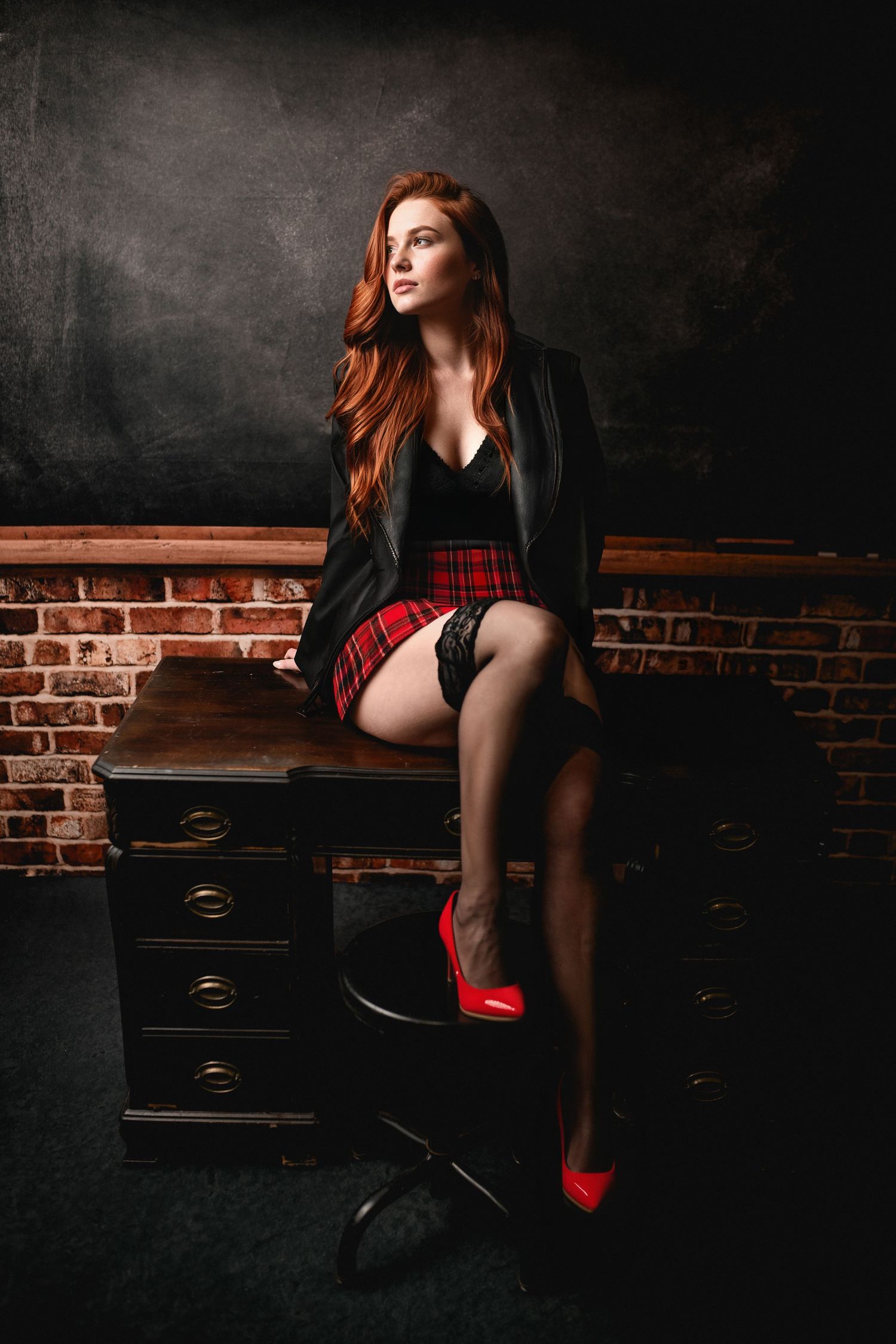 Redhead woman in plaid skirt, black blazer, and red heels sitting on vintage desk against brick wall.