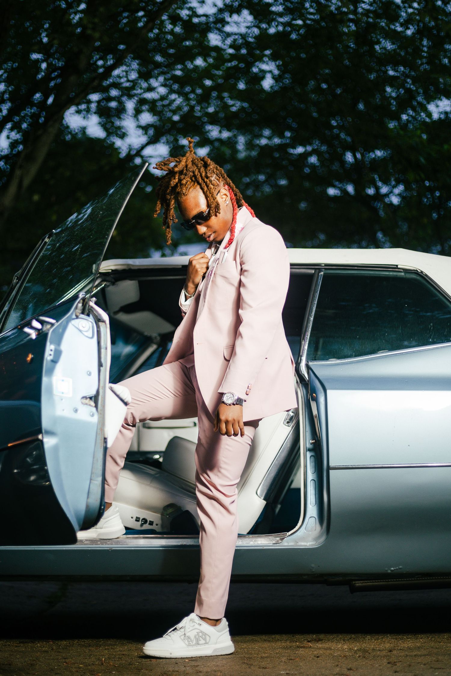 A stylish young man in a pink suit with dreadlocks poses stepping out of a silver vintage car outdoors.