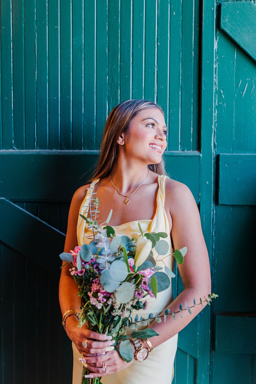 Smiling woman in yellow dress holding a bouquet of wildflowers against a teal green wooden wall.