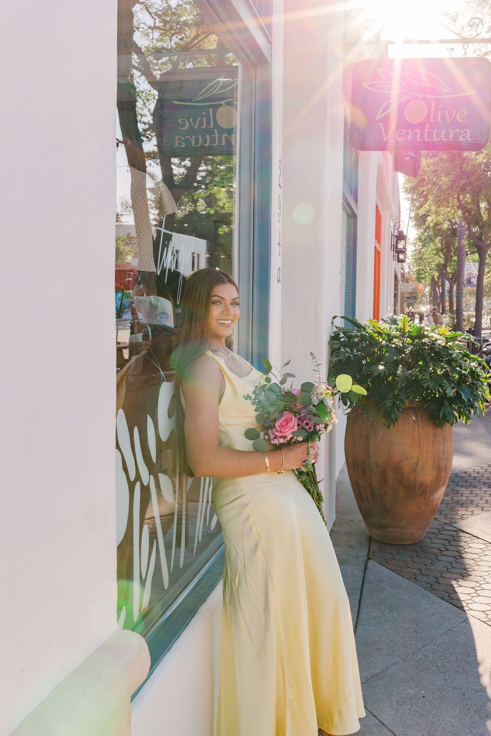 Woman in yellow dress holding bouquet poses outside Olive Ventura shop in golden sunlight.