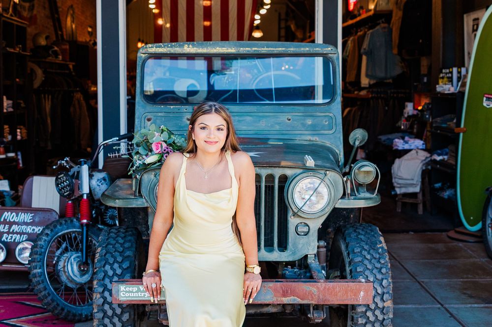 Woman in yellow slip dress posing in front of a vintage green Jeep outside a rustic shop.