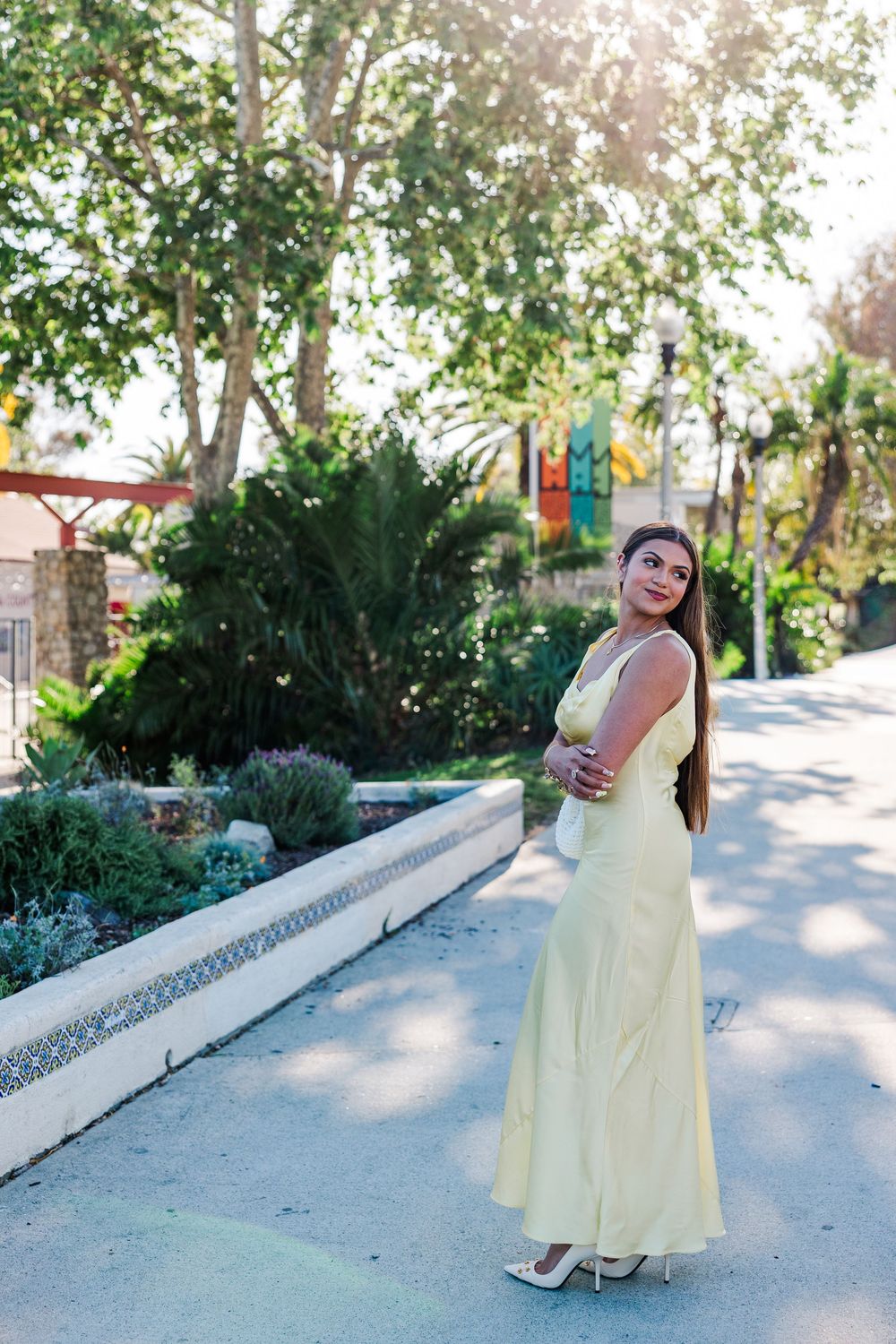 Young woman in elegant yellow maxi dress posing outdoors in a lush garden pathway on a sunny day.