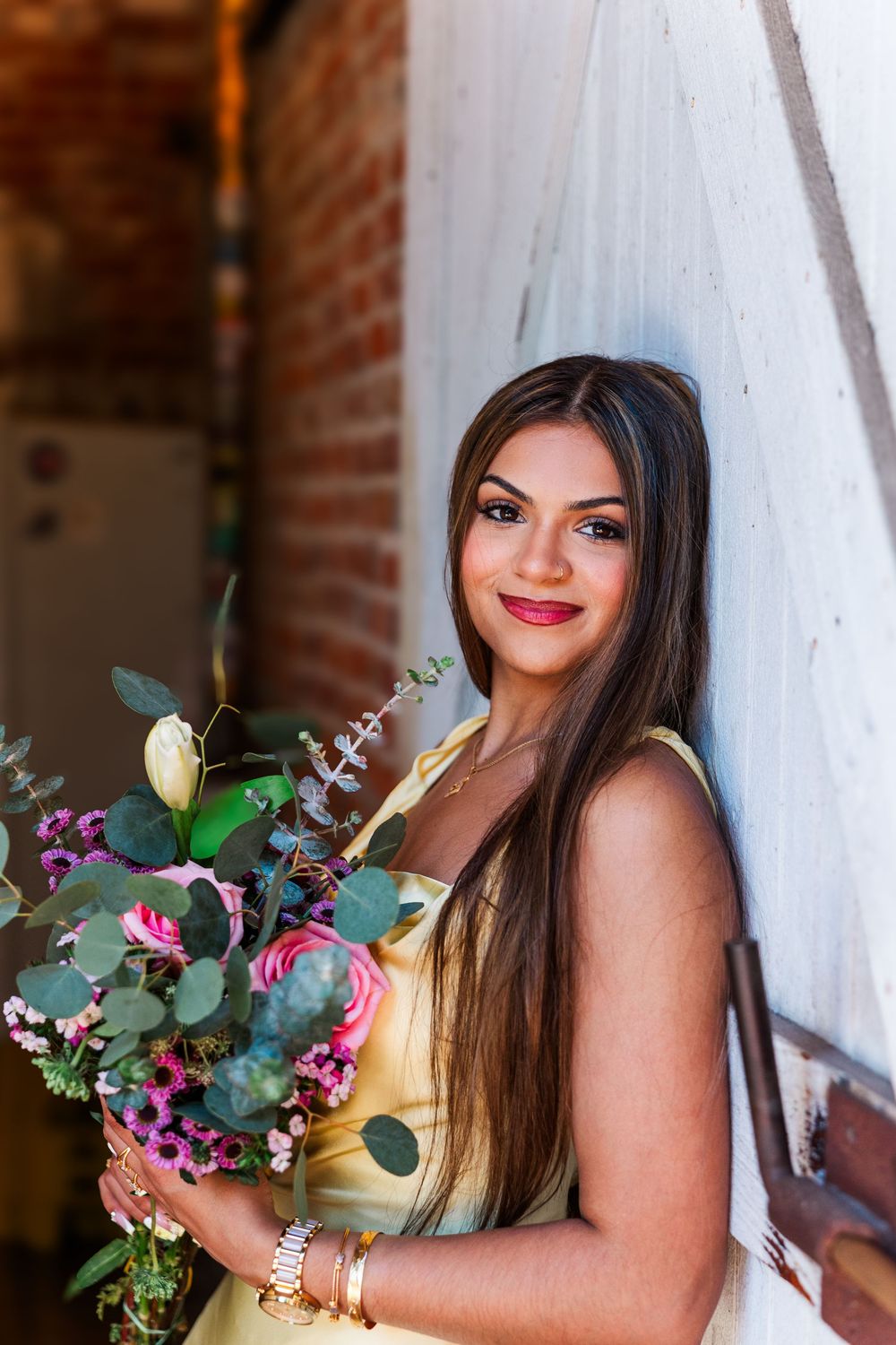 Young woman in yellow dress holding colorful bouquet of flowers, leaning against wall, smiling.