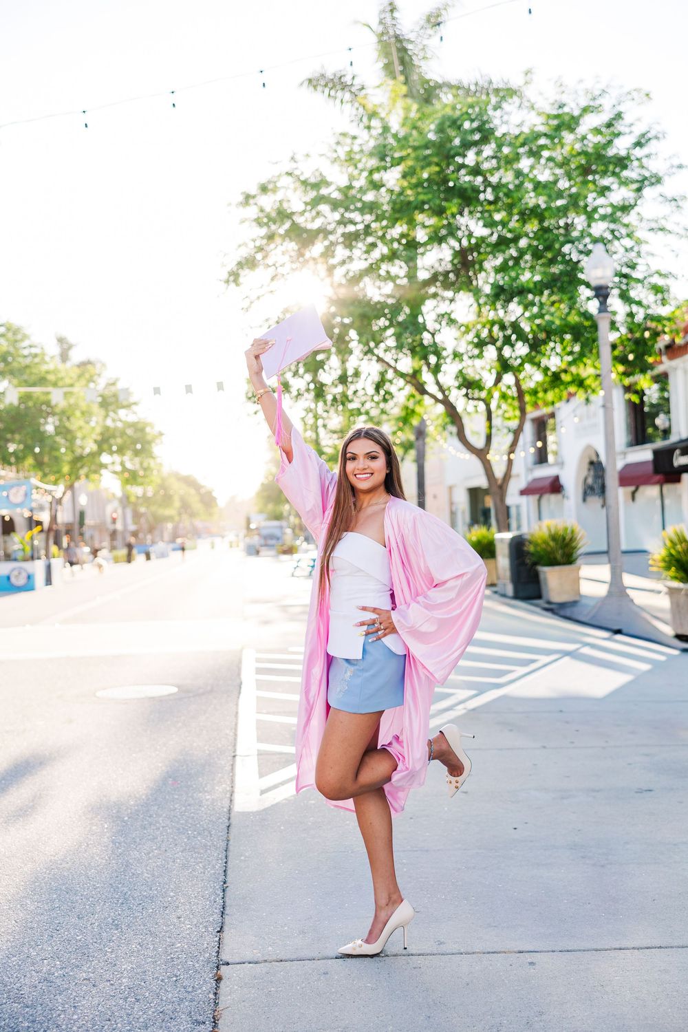 Young woman in pink graduation robe raises diploma on sunny city sidewalk, celebrating her academic achievement.