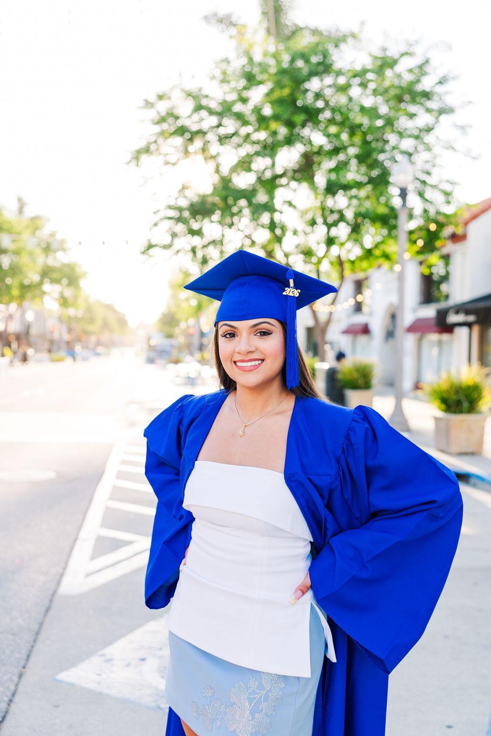Smiling young woman in blue graduation cap and gown posing outdoors on a sunny street after her graduation ceremony.