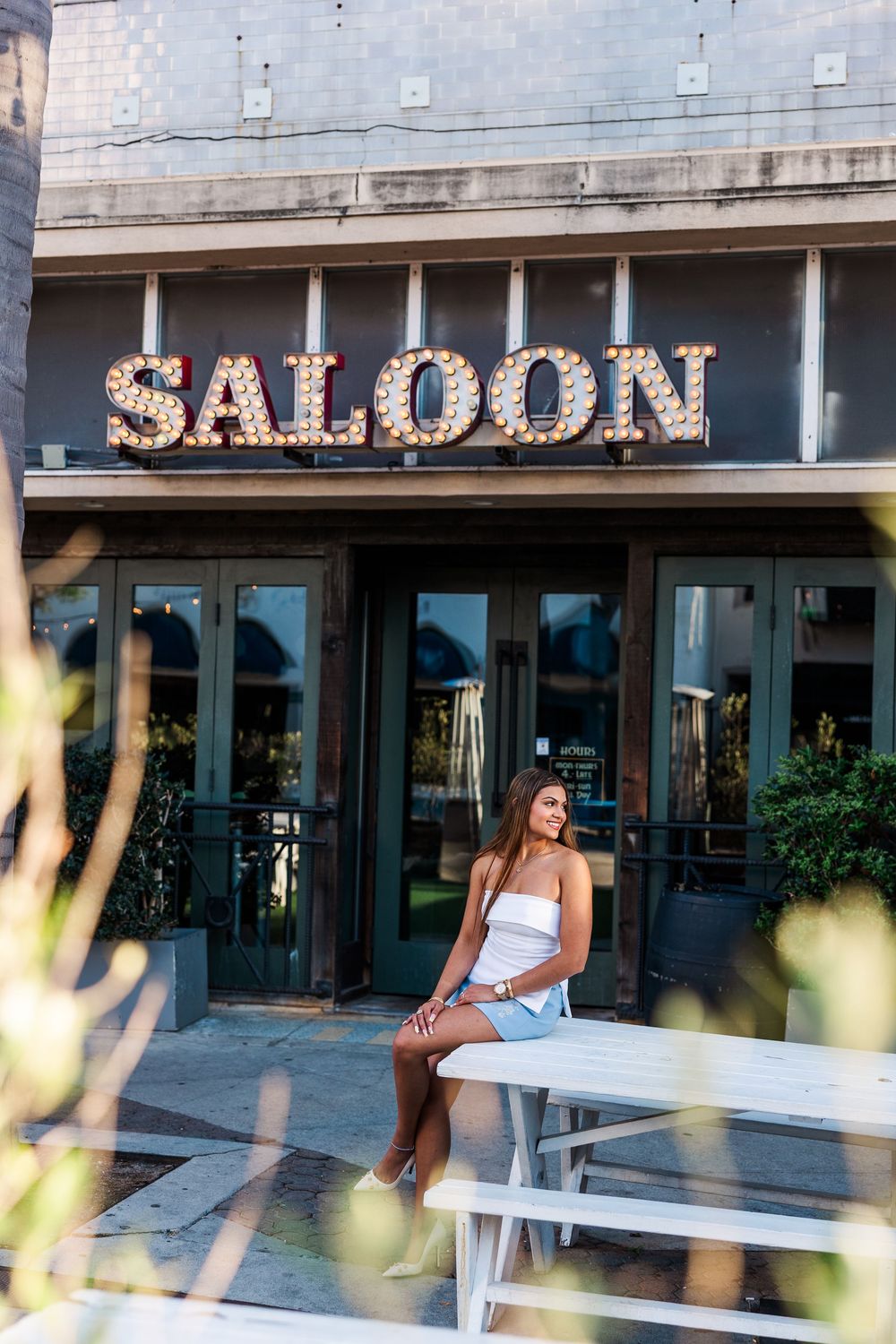 Young woman in white top and denim skirt sitting outside a Saloon bar with marquee lights.