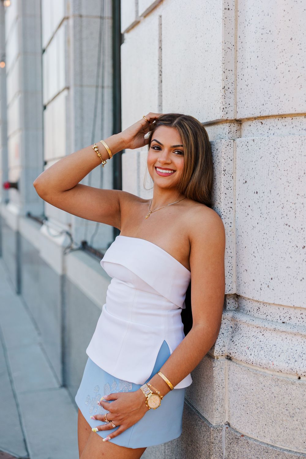 Smiling young woman in white strapless top and blue skirt leaning against a stone wall wearing gold jewelry.
