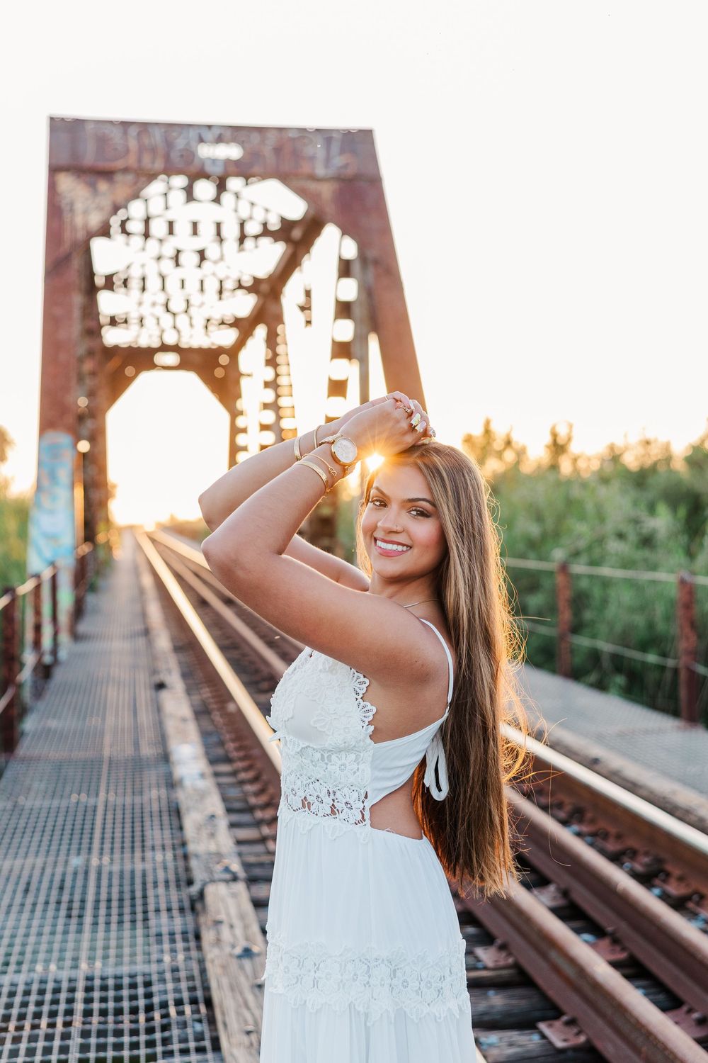 Smiling woman in white lace dress on railway bridge at golden sunset hour.