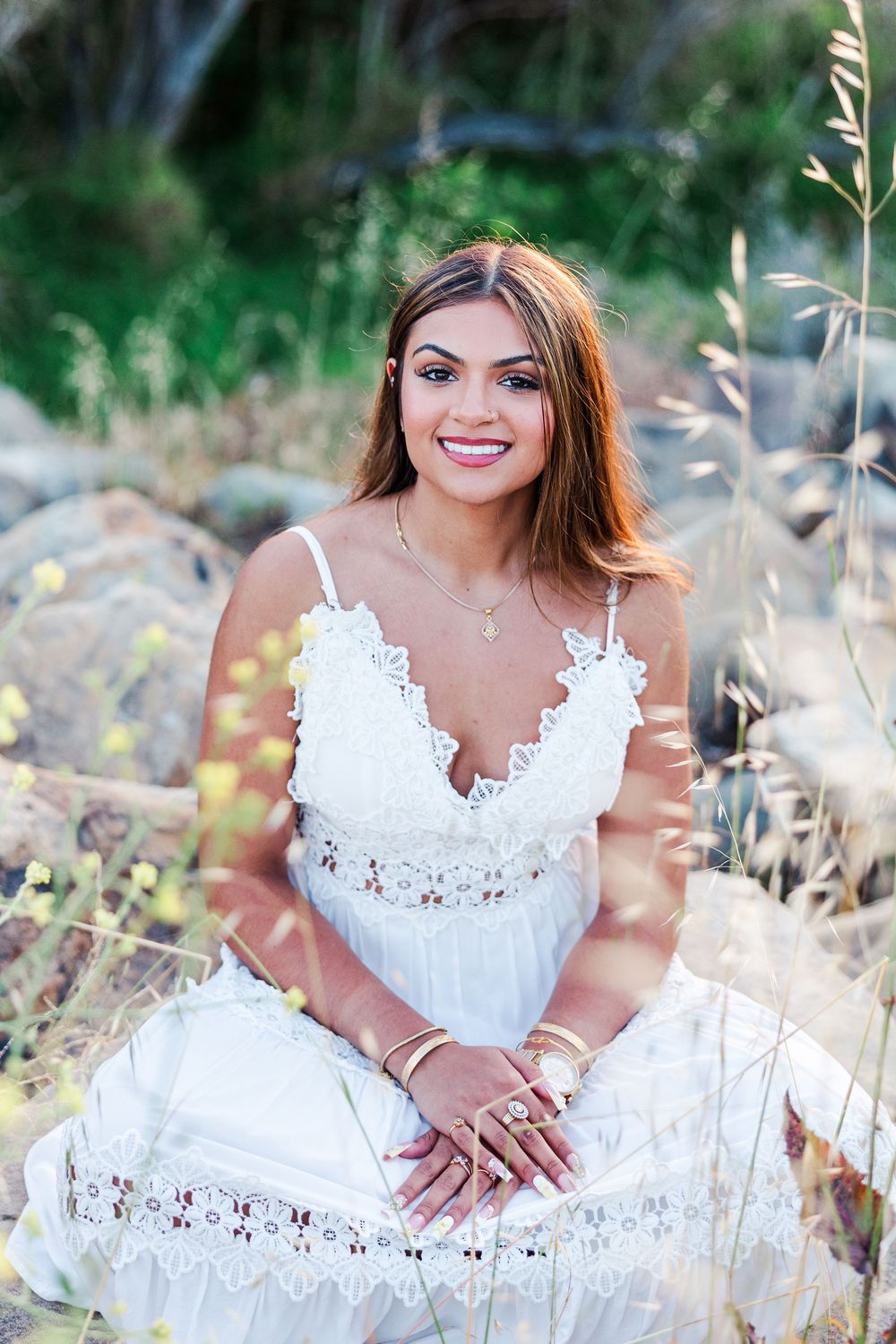 Smiling young woman in white lace dress sitting among tall grass and rocks outdoors in natural light.