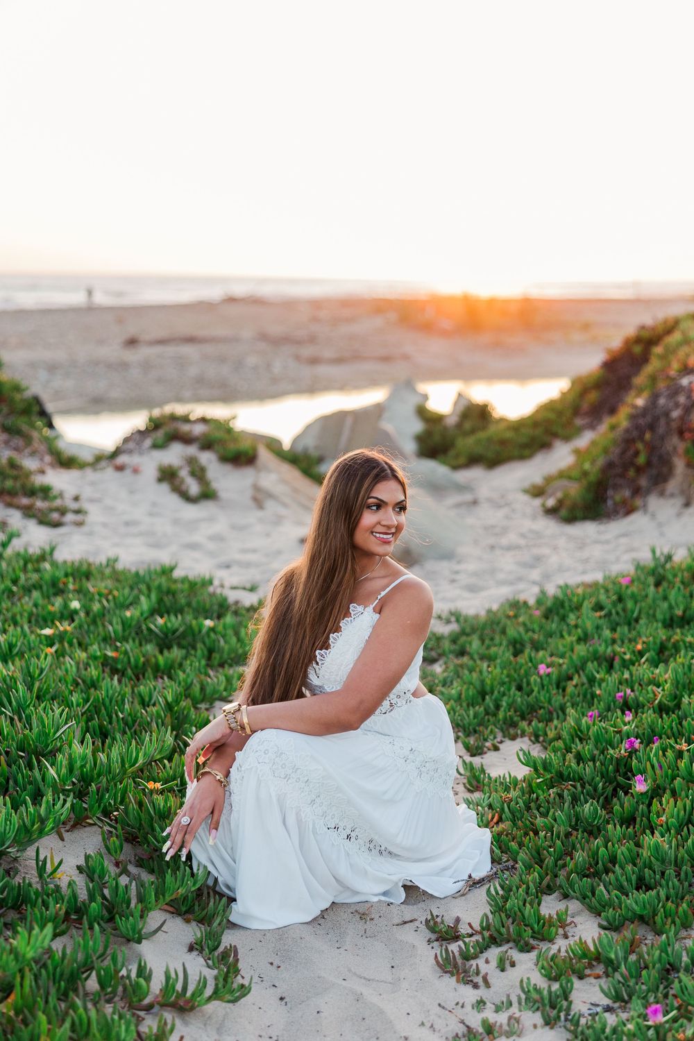 Young woman in white dress sitting on sandy beach dunes with green plants at golden sunset.