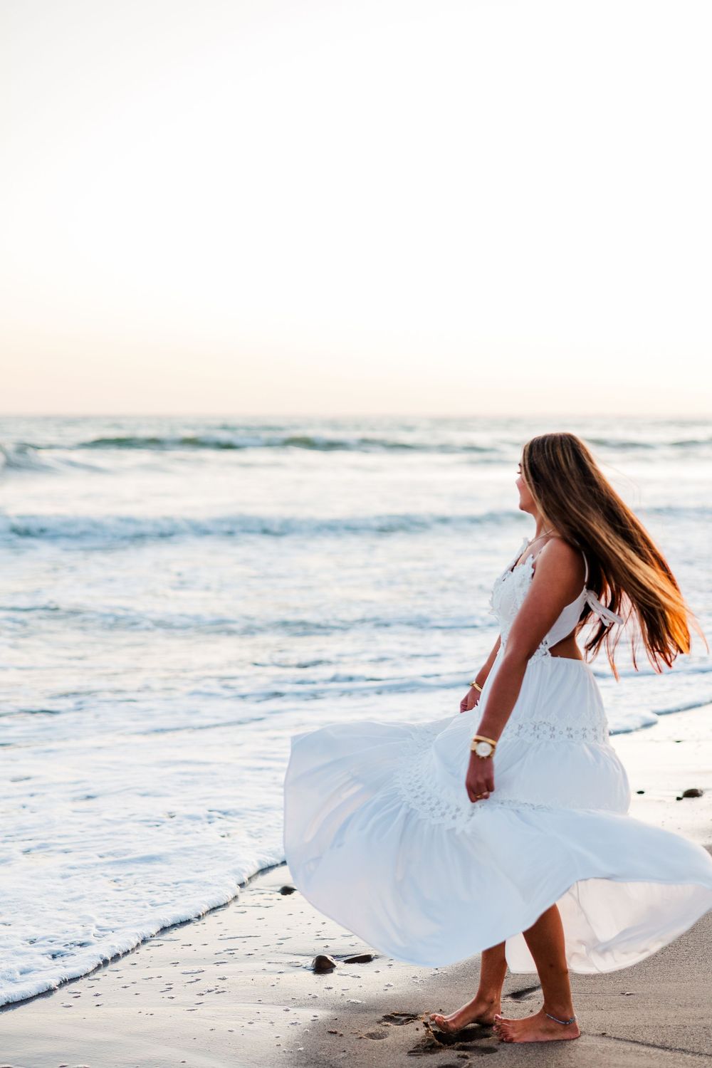 Woman in flowing white dress standing on beach at sunset with ocean waves.
