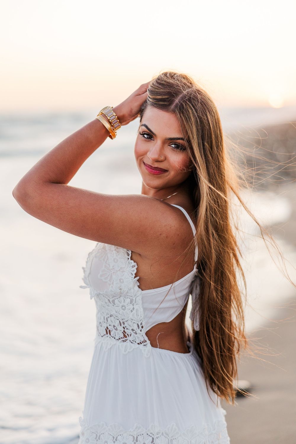 Young woman in white lace dress smiling at beach sunset, long hair blowing in wind, gold bracelet accessory.