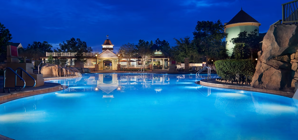 Illuminated resort swimming pool at night with glowing blue water and surrounding buildings.