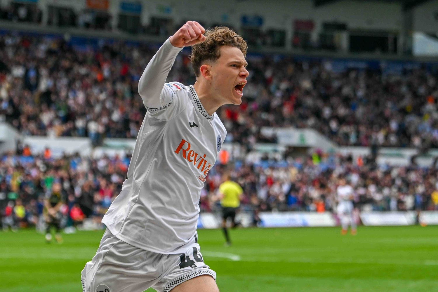 Young footballer celebrates a goal in a packed stadium, wearing a white jersey with red sponsor logo.